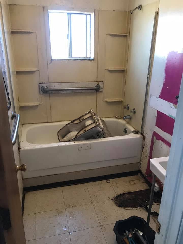 Small bathroom with a tub, window, shelves, and a cluttered floor; a bag sits in the tub.