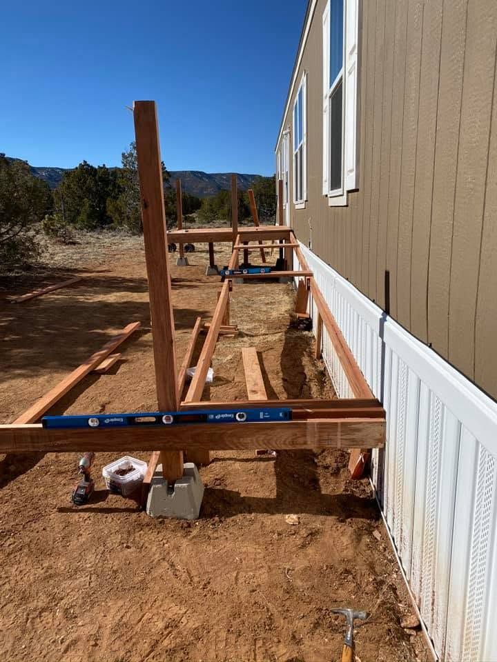 House foundation under construction beside tan siding, with wooden posts, concrete footings, and a blue level.
