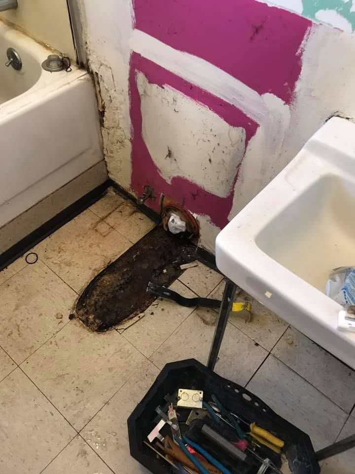 Bathroom corner with exposed wall and floor damage beside a sink and bathtub, with tools in a tray.