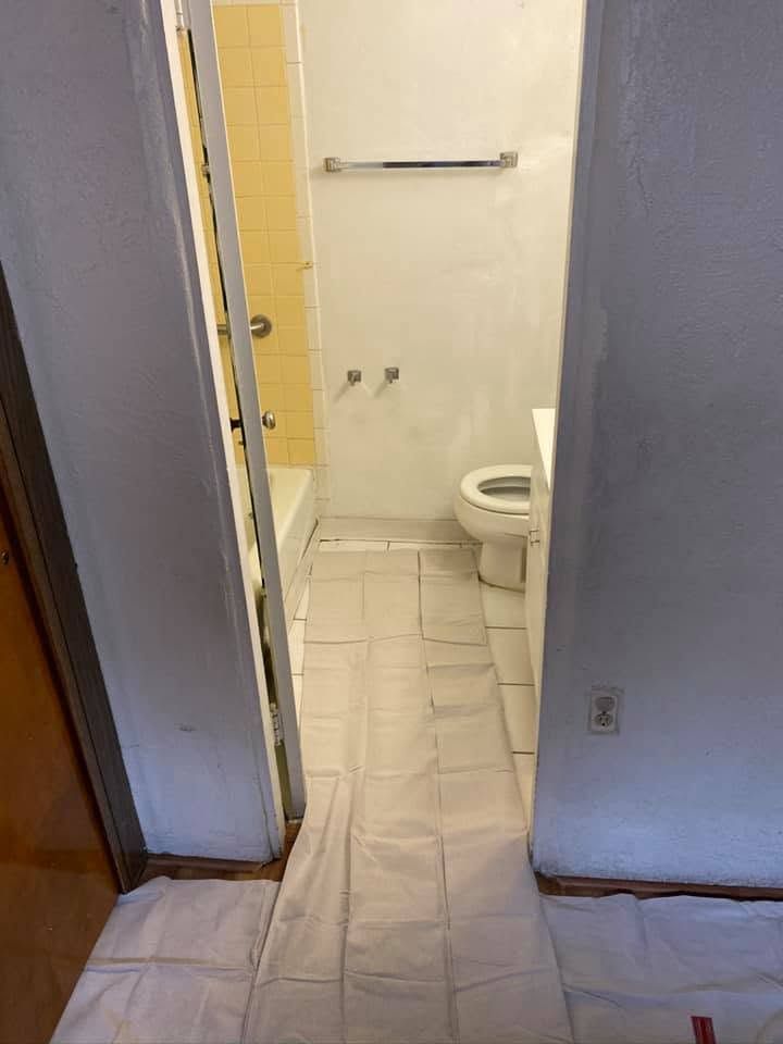 Narrow bathroom hallway with white tile floor, open door, towel bar, and toilet visible in the back.