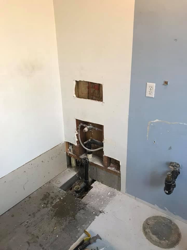 Damaged wall with exposed plumbing and open cutout in a room, near a gray wall and floor drain.