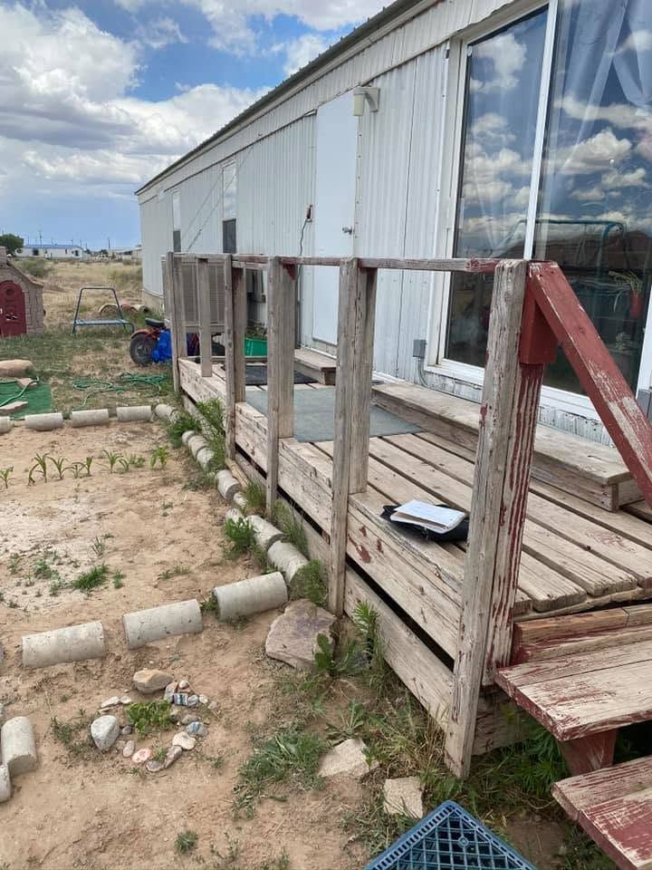 Weathered wooden porch with red stairs beside a white mobile home on a rocky dirt lot.