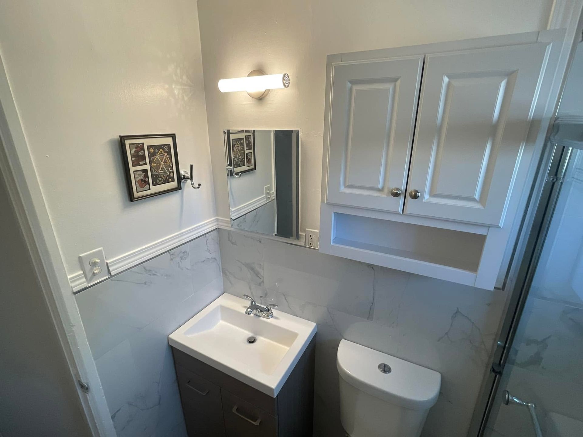 Small bathroom with white vanity, toilet, mirror, wall cabinet, and shower edge.
