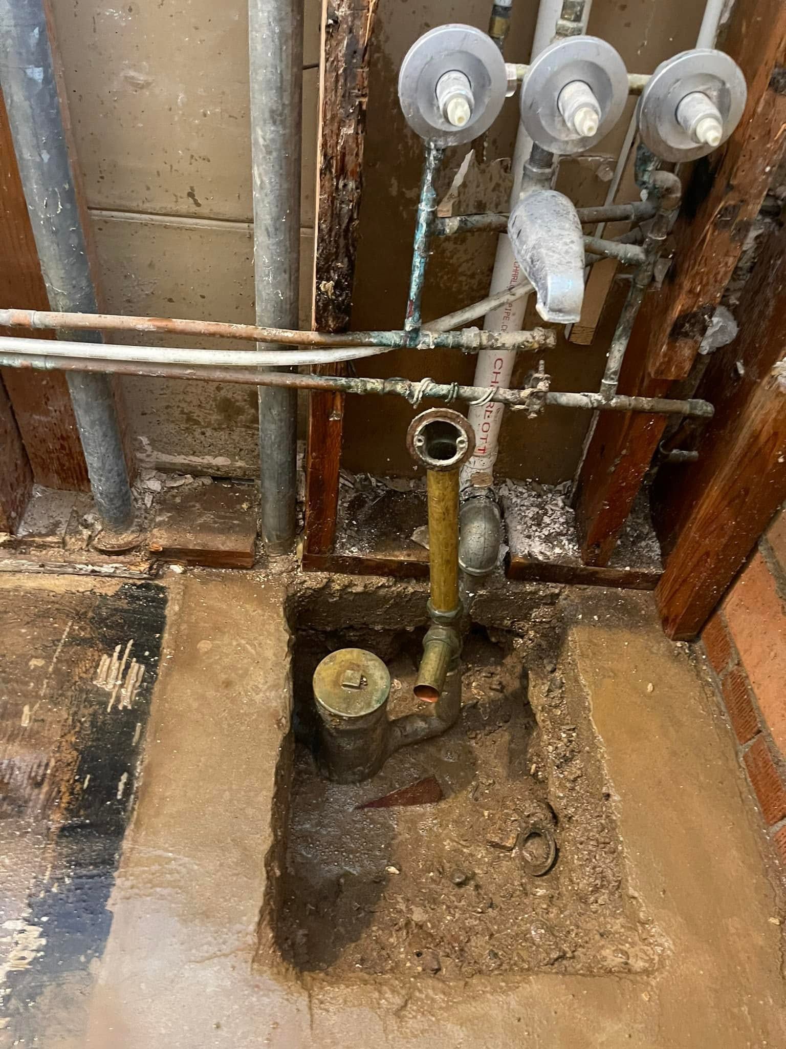 Basement utility area with exposed pipes, meters, and a floor drain amid damp, unfinished concrete.