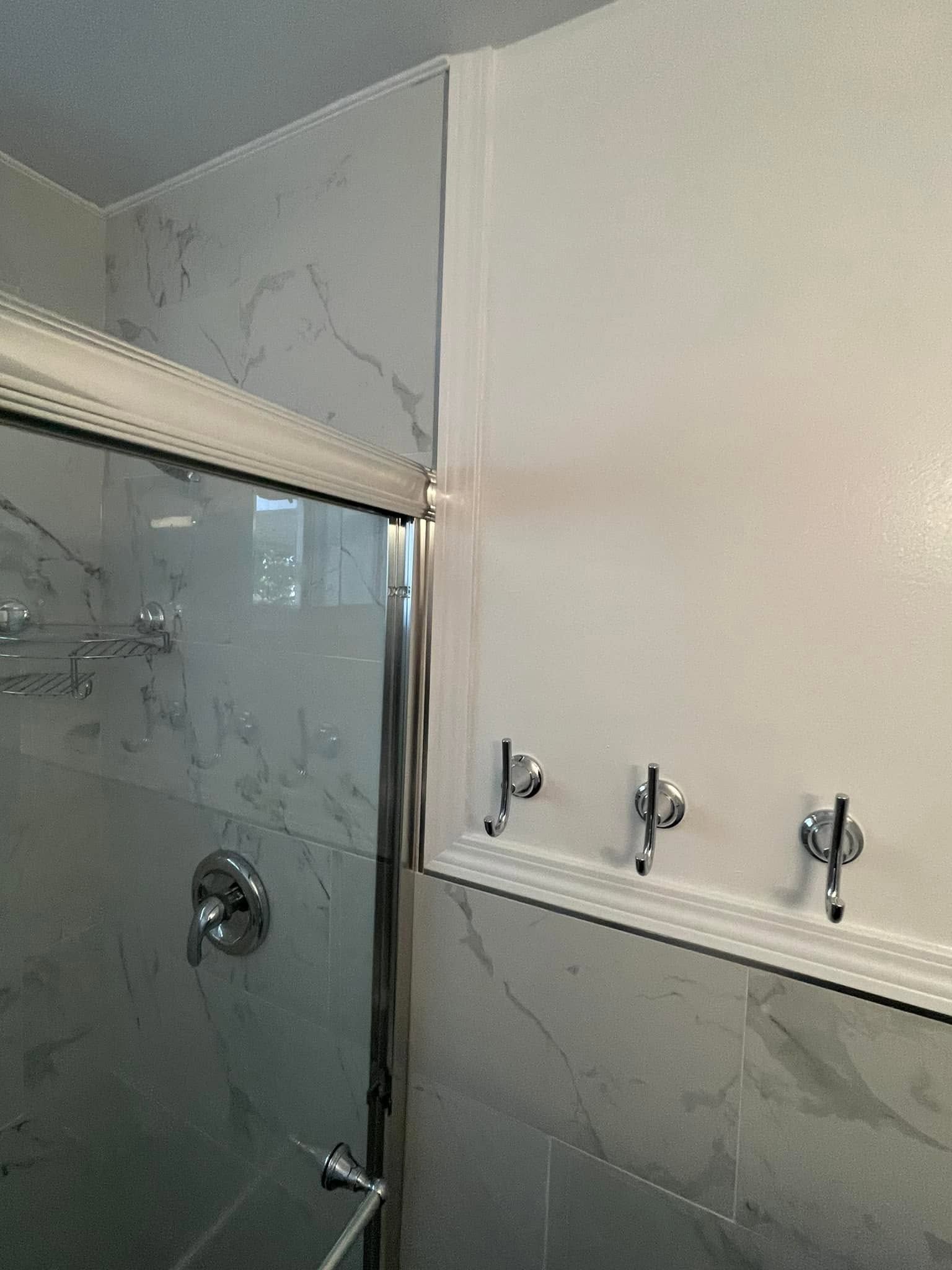 Bathroom corner with marble tile shower, glass door, and white wall with three hooks showing water damage at the ceiling edge