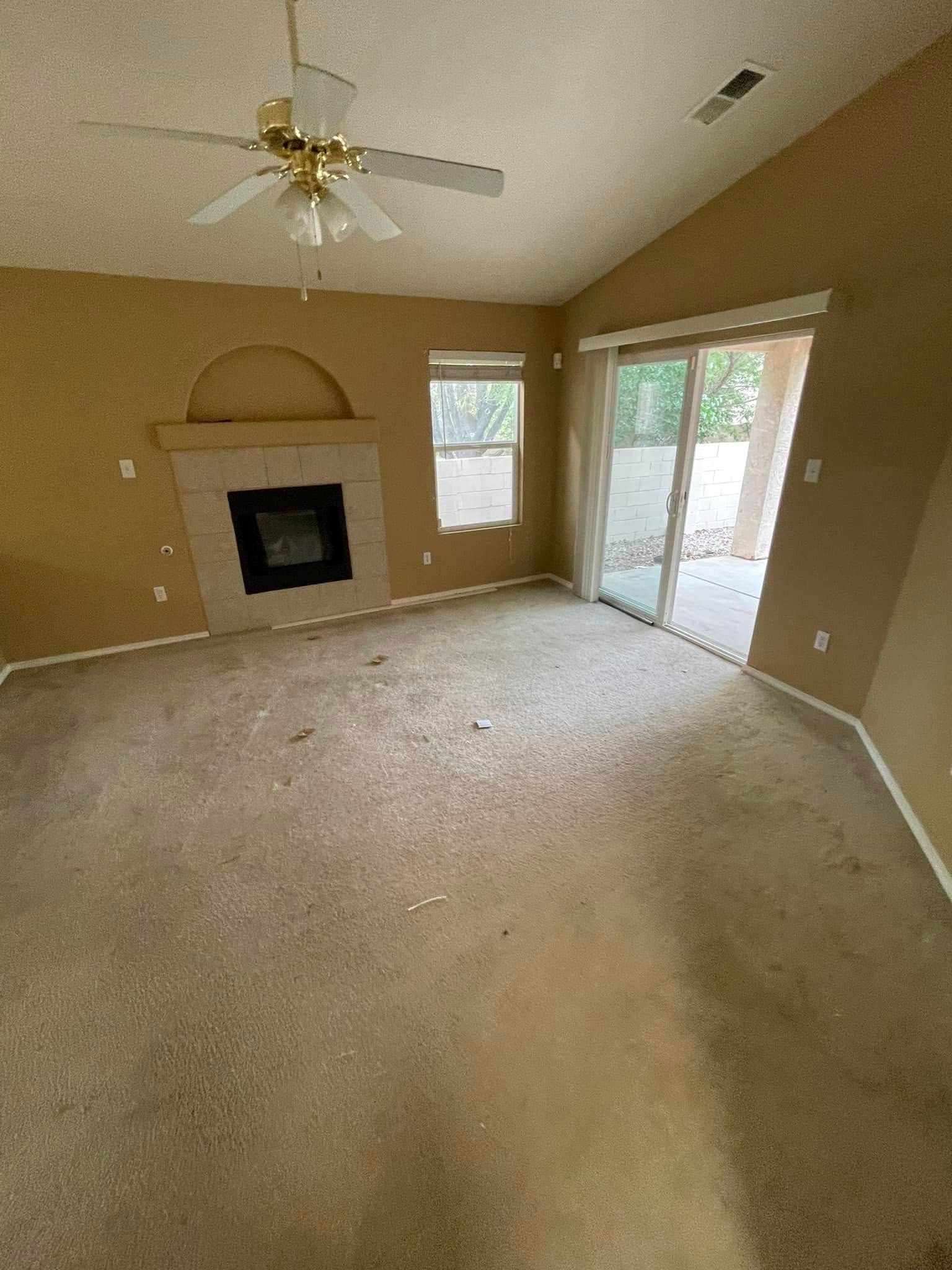 Empty beige living room with fireplace, ceiling fan, carpet, and sliding glass door to outside.