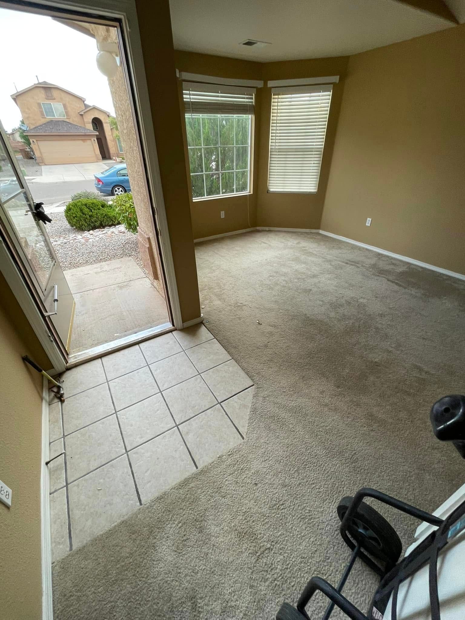 Empty carpeted room with tiled entryway, front door open to a street, and a window with blinds.
