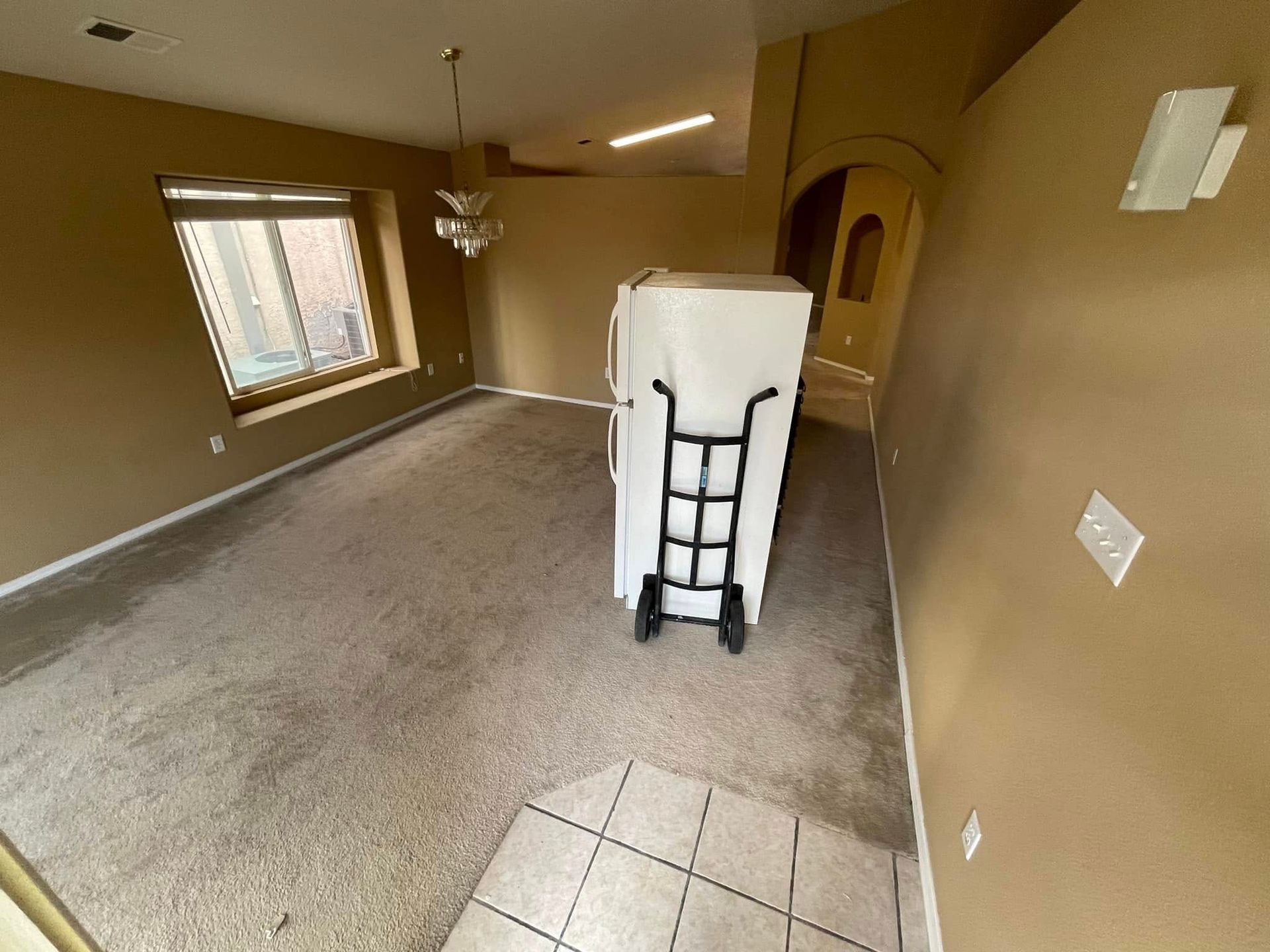 Empty tan living room with carpet, tile entry, ceiling fan, and a covered item on a dolly in the center.