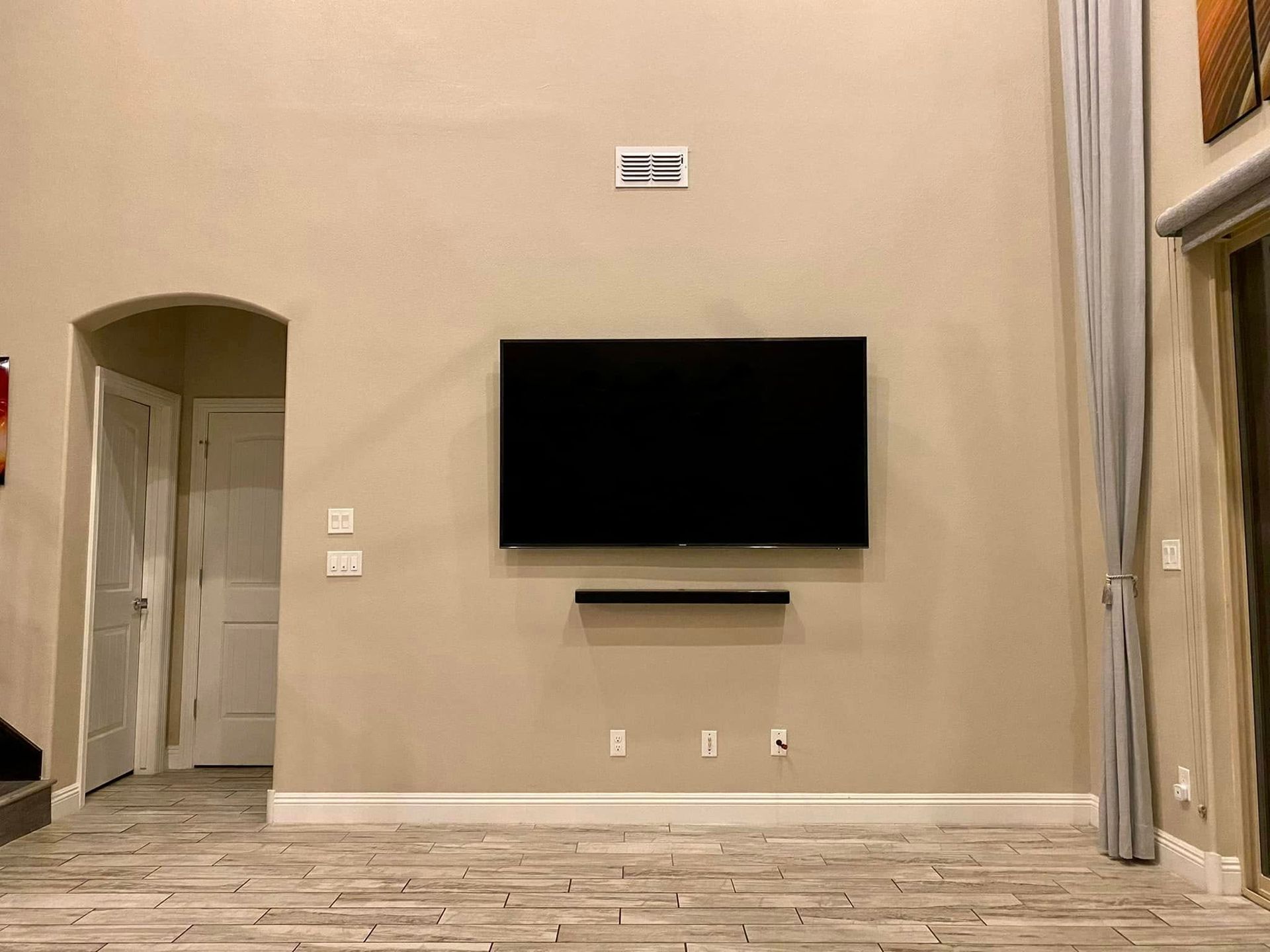 Empty living room with beige walls, wall-mounted TV, and arched doorway, viewed from a tiled floor.