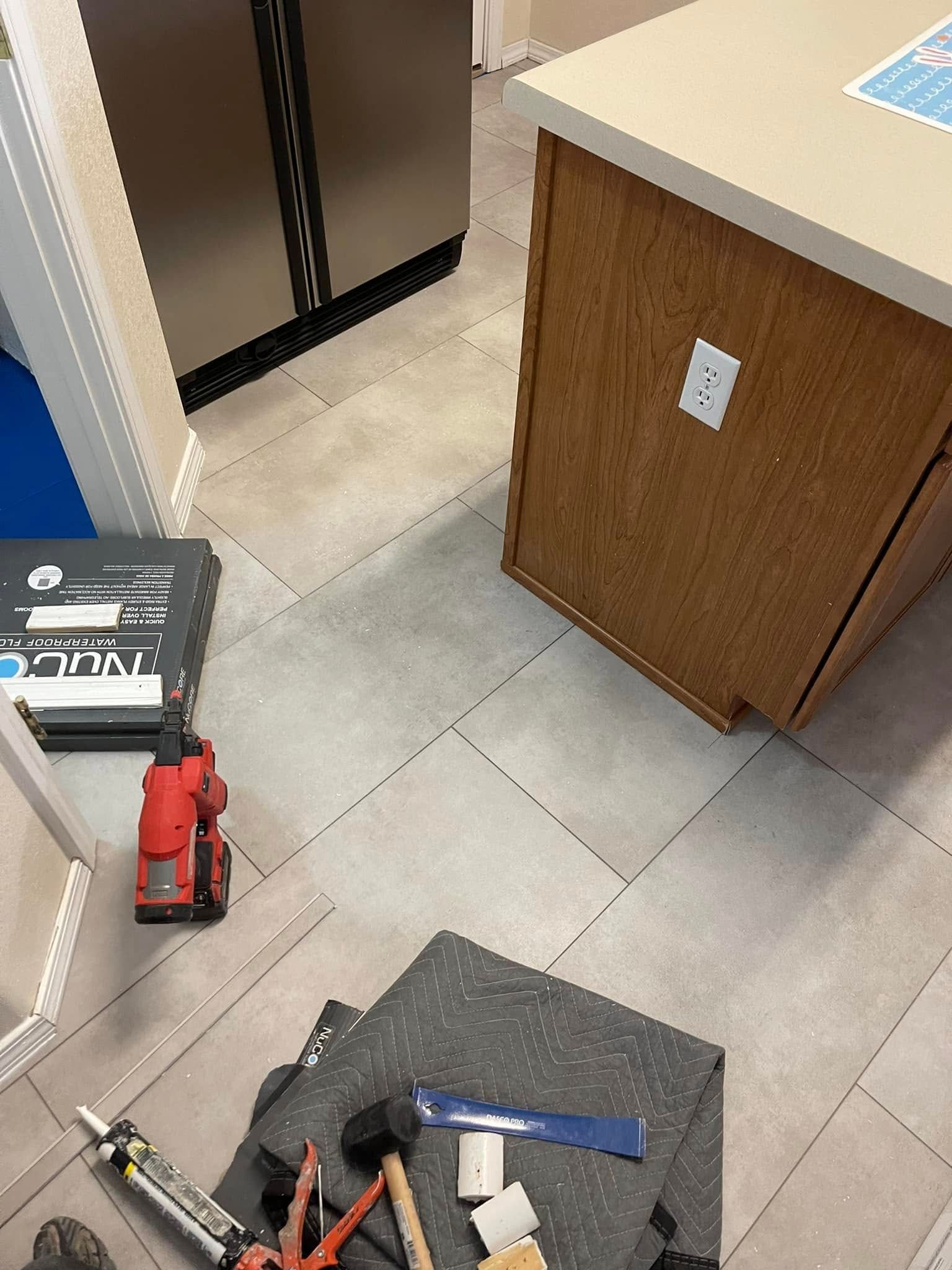Cluttered garage corner with tools, a black cart, red jack, and wooden cabinet on tiled floor.