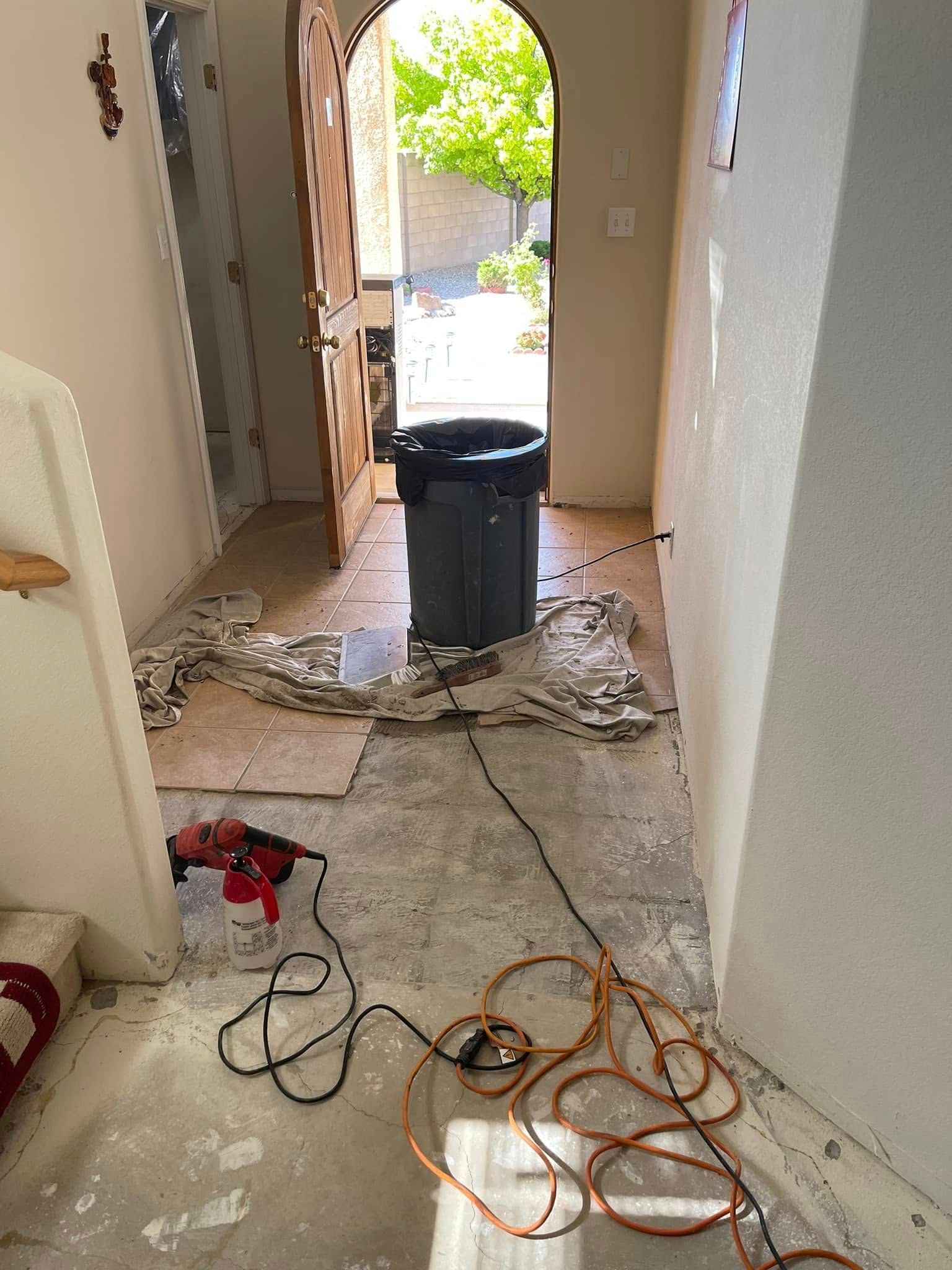 Damaged hallway with muddy floor, construction debris, cords, and a trash can near an open front door