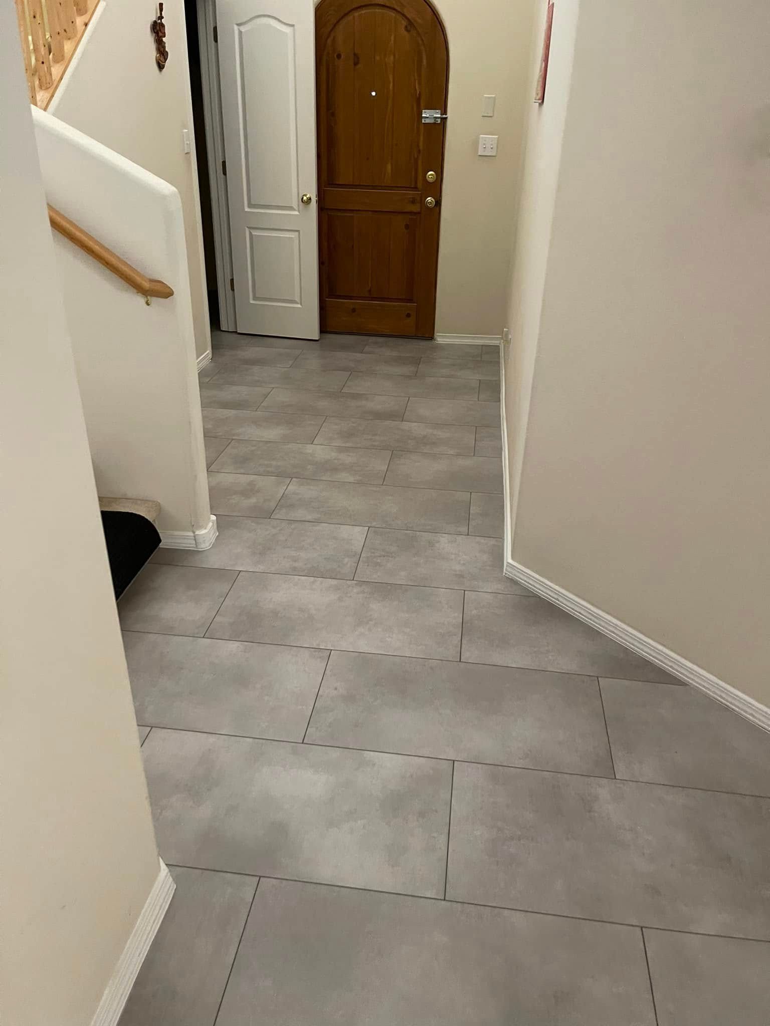 Narrow hallway with gray floor tiles, white walls, stairs on left, and a wooden front door ahead.