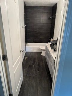 Narrow bathroom with dark wood floor, white walls, tub-shower at the back, and a toilet beside a countertop.