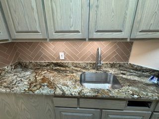 Kitchen countertop with granite surface, stainless sink, wood cabinets, and herringbone tile backsplash
