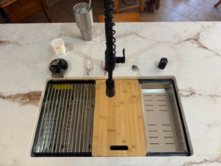 Kitchen sink with wooden cutting board over basin, faucet, dish rack, and mugs on a marble counter.