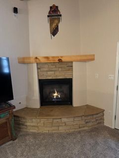 Corner fireplace with stone surround, wooden mantel, and a small fire burning in a carpeted living room.