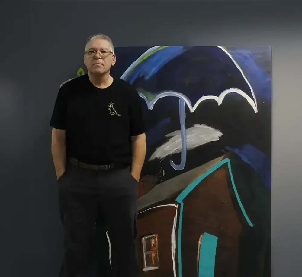 Martin Smith standing in front of a painting by Ed Besh showing a house with an umbrella floating above it.