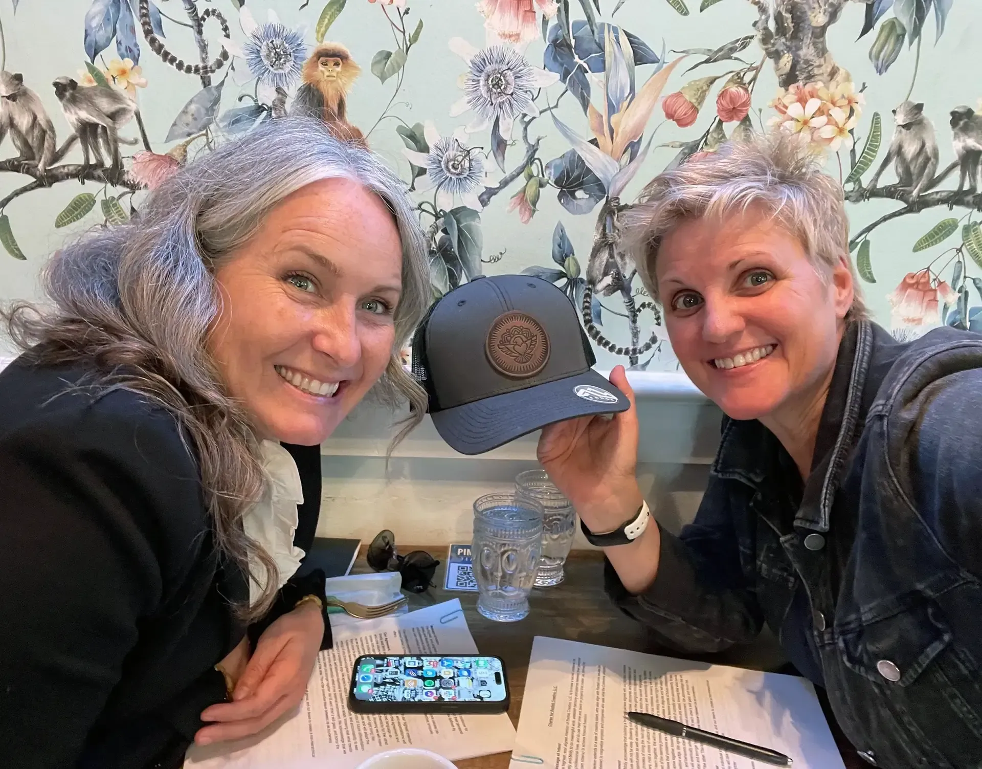 Two women are sitting at a table holding a hat and smiling