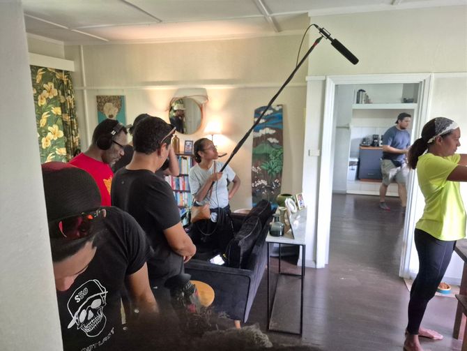 Filming a scene indoors. Crew members surround a woman; a boom mic hovers. Others stand in the kitchen doorway.