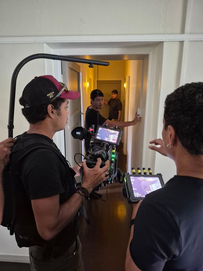 Film crew filming in a hallway. One operates camera, two watch monitors, and one actor waits.
