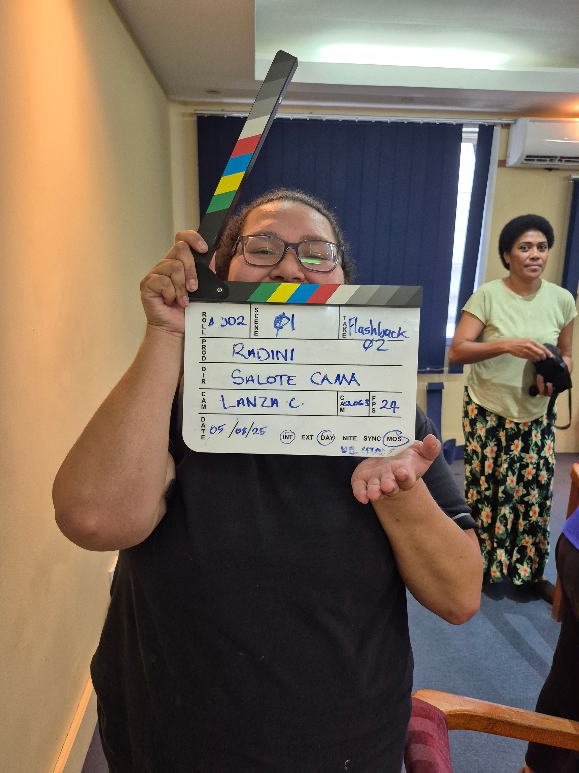 Person holding a film clapperboard, blowing a kiss. Another person stands behind in a room.
