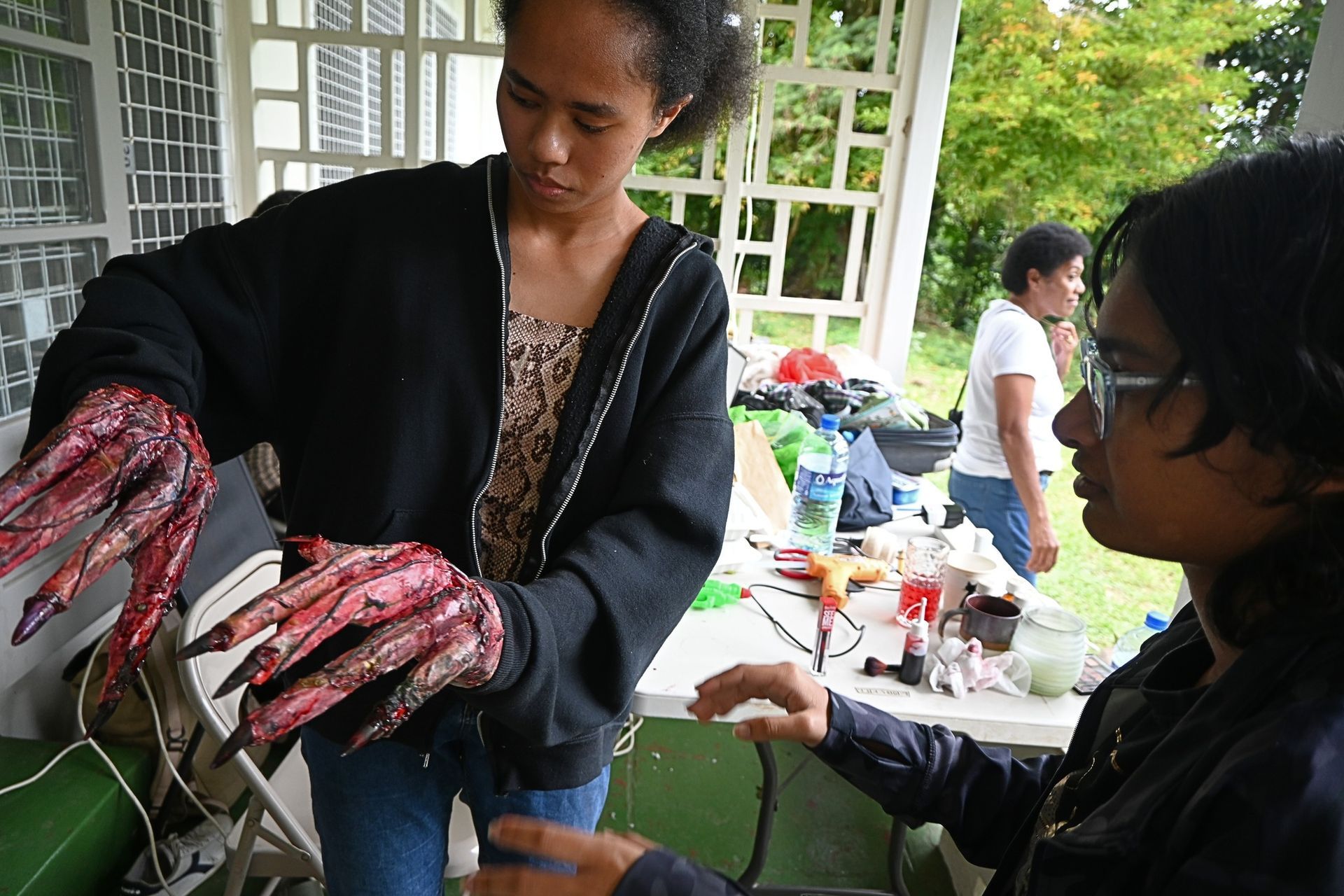 Two people examine prosthetic, bloody claws; outdoor setting with others working in background.