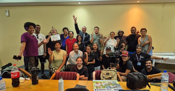 Group of diverse people smiling and waving in a room, some wearing casual clothes, gathered around a table.