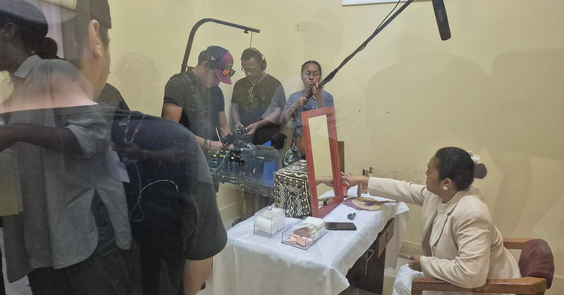 A film set with people directing an actress at a vanity table. The actress is pointing at something.