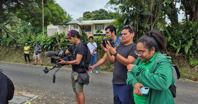 A film crew records on a road. People hold cameras; a building and foliage are in the background.