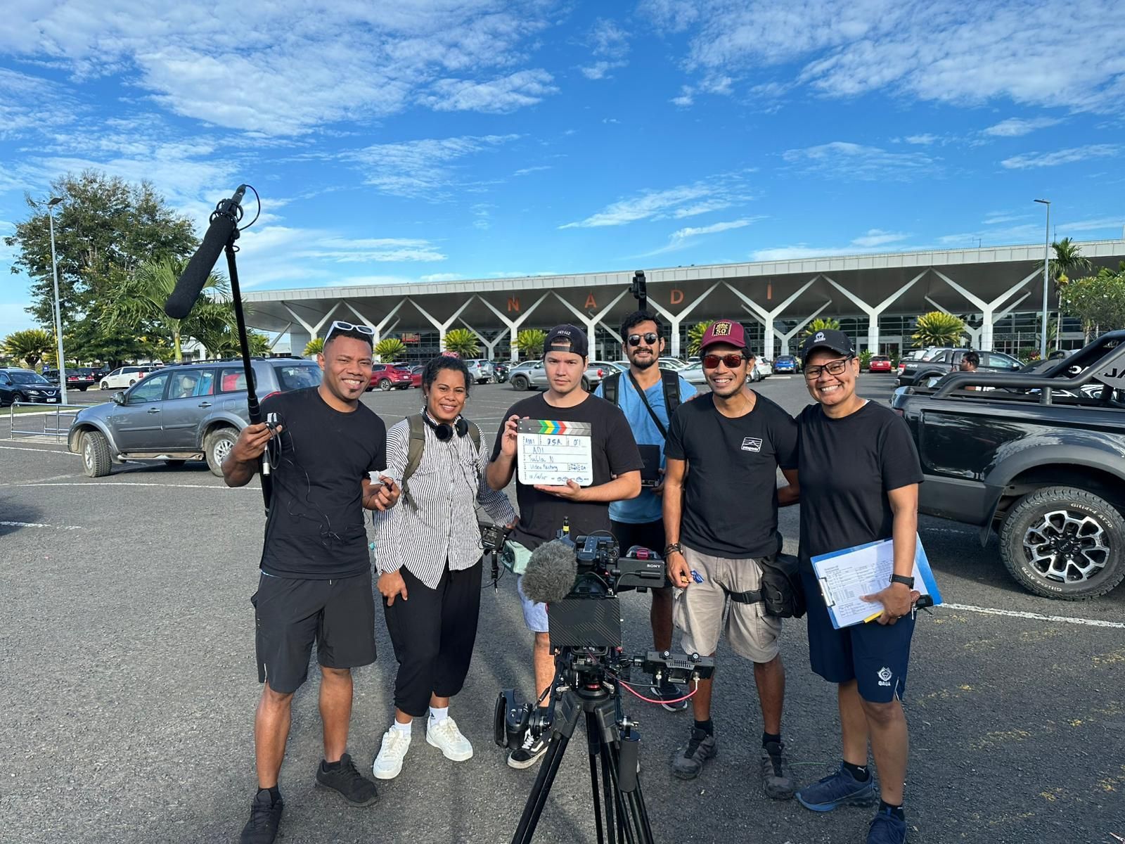 Film crew posing outdoors near a building. Crew holds clapperboard, microphone, and camera. Sunny day.