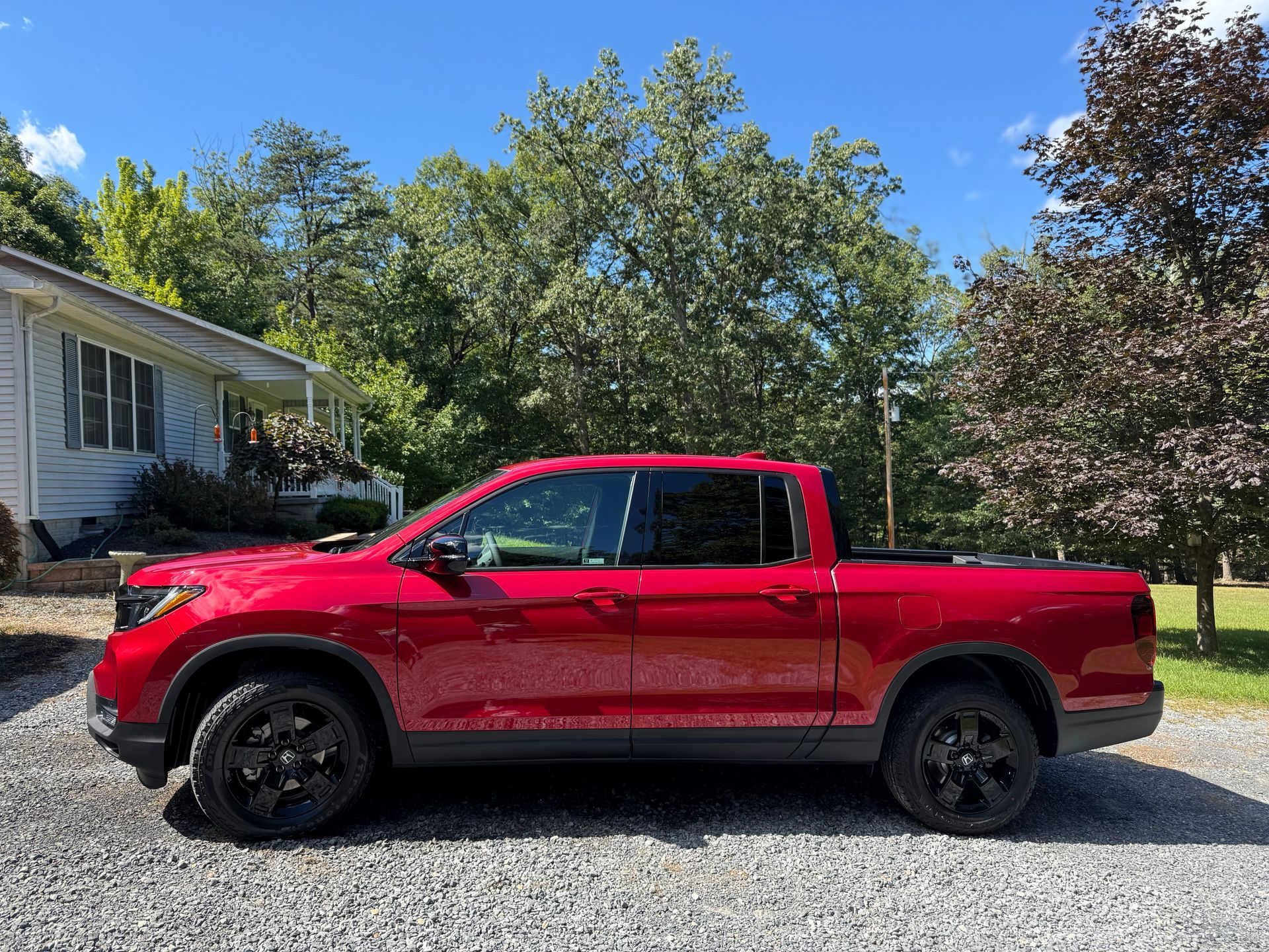 Red Honda Ridgeline truck parked on a gravel driveway, with a house and trees in the background.
