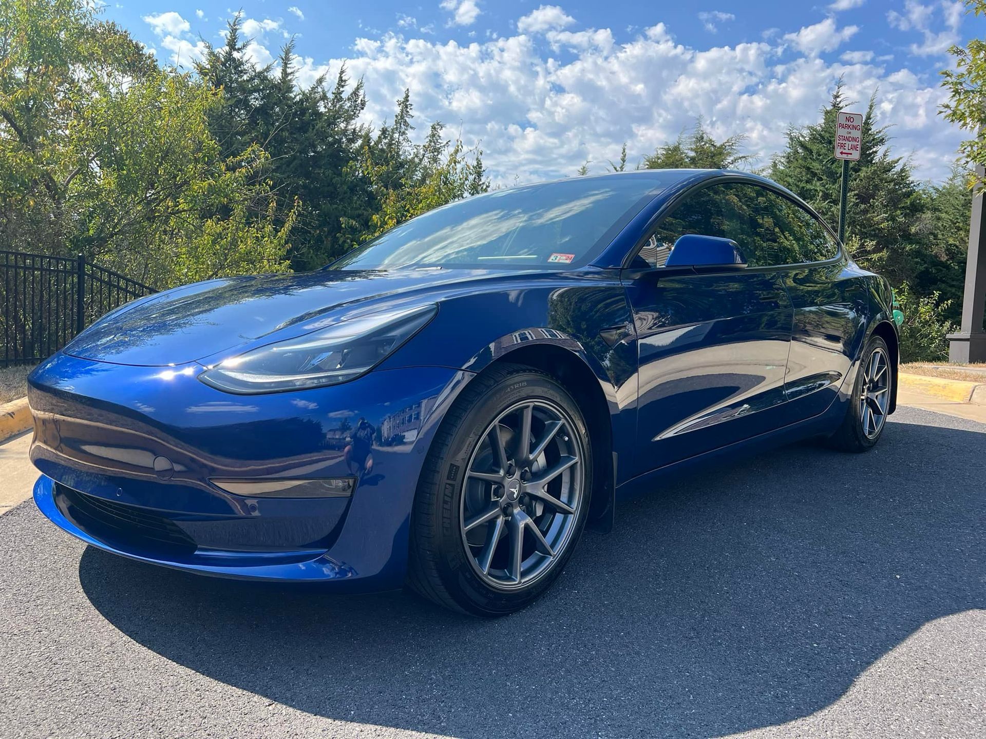 Blue Tesla Model 3 parked on asphalt in front of a building with trees and blue sky.