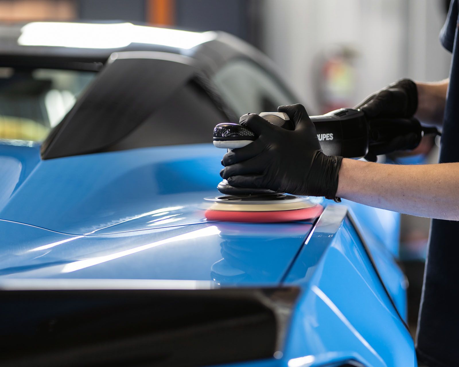 Person polishing blue car with electric polisher, wearing black gloves.
