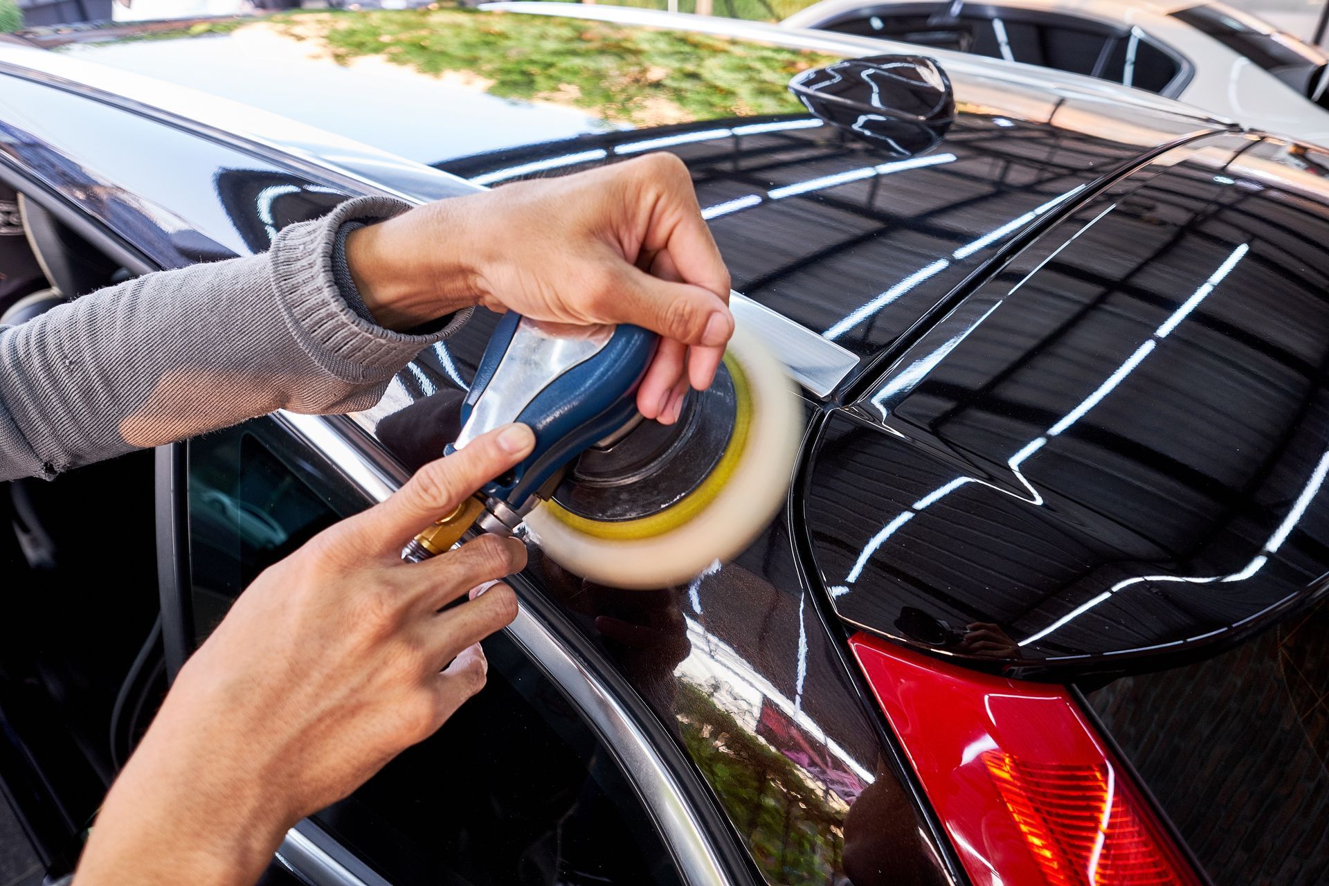 Person polishing a black car with a buffer; close-up shows hands holding the machine.