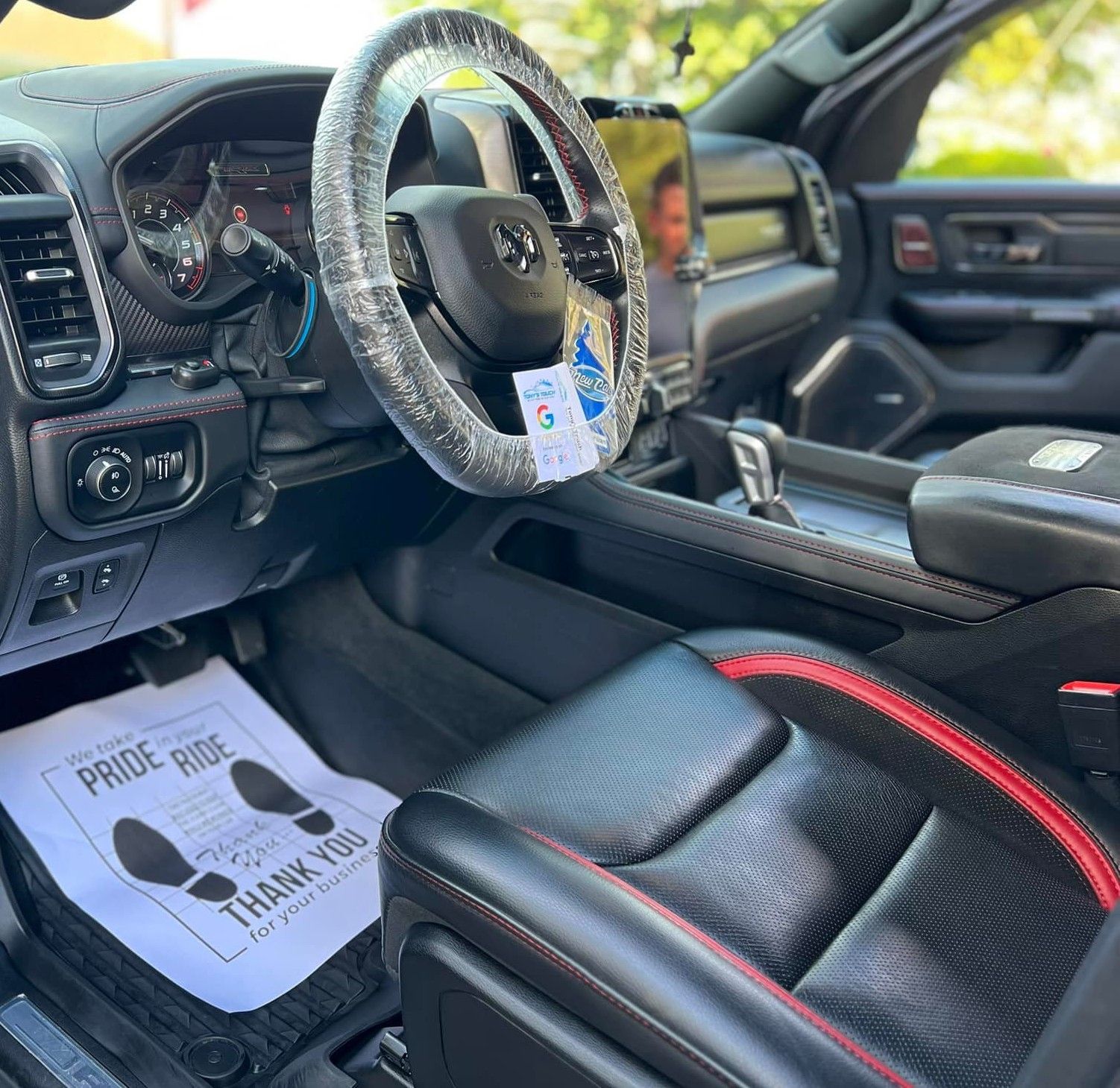 Interior of a black RAM truck; black leather seats with red trim, steering wheel wrapped in plastic, paper floor mat.