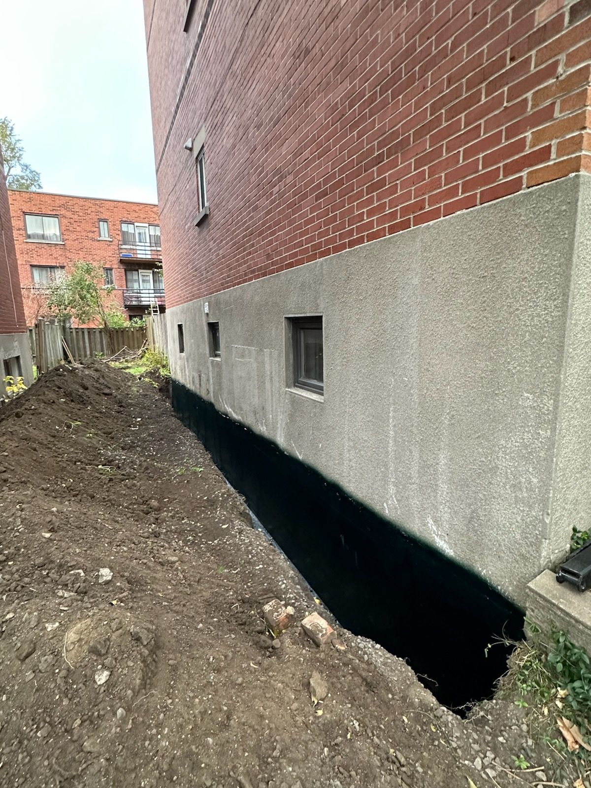 Tranchée creusée le long des fondations d'un bâtiment en briques, laissant apparaître une membrane d'étanchéité noire.
