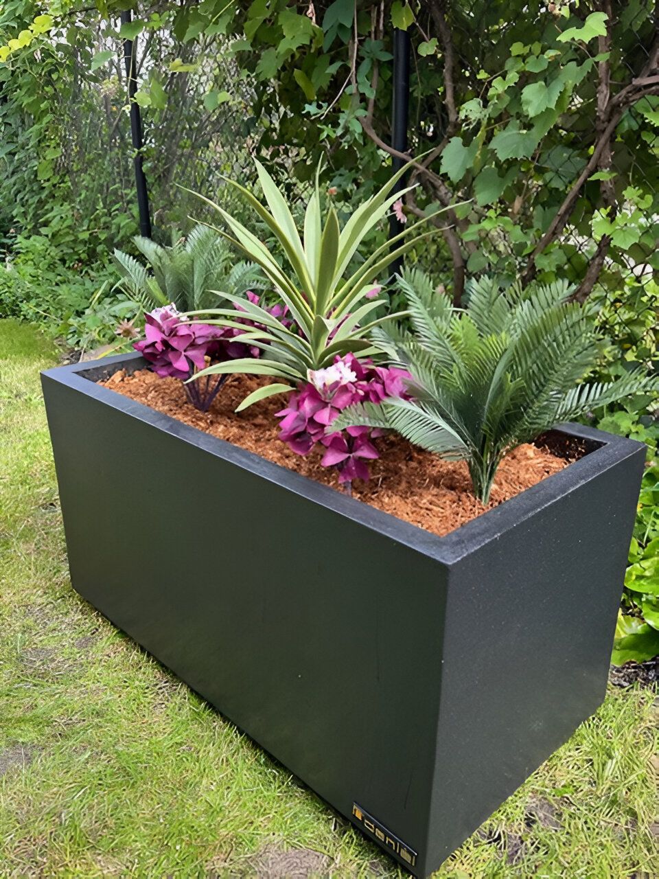 Une jardinière noire remplie de fleurs et de plantes est posée dans l'herbe.