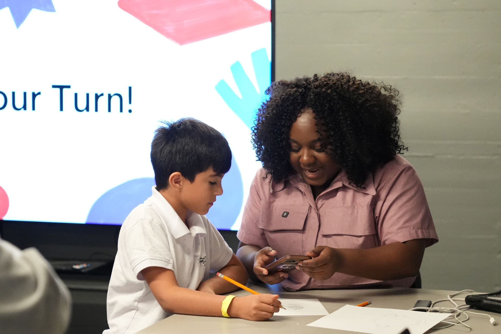 Boy wearing headphones, using a laptop at a wooden table, notebook nearby. Kitchen in background.