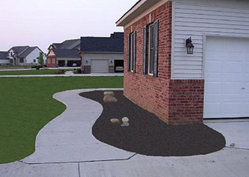 Curved concrete walkway beside a brick house, bordered by dark mulch and small rocks in a suburban yard