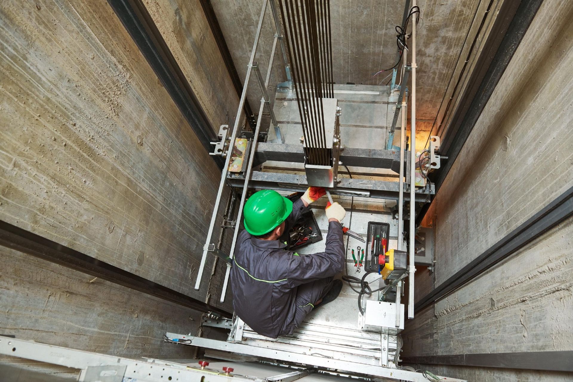 Worker in green hard hat on a suspended lift inside a tall industrial shaft, using equipment.