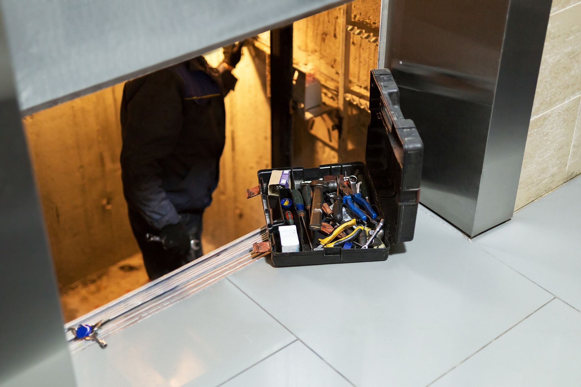 Person crouching under a large machine beside an open electronics chassis in a tiled indoor area