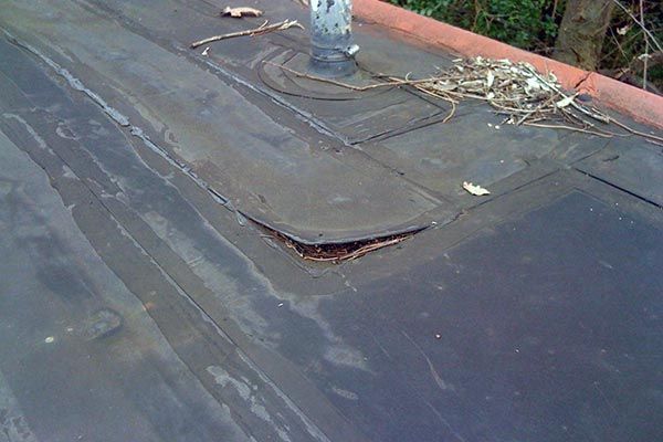 A damaged section of a black flat roof with a peeling edge and a pipe vent surrounded by debris.