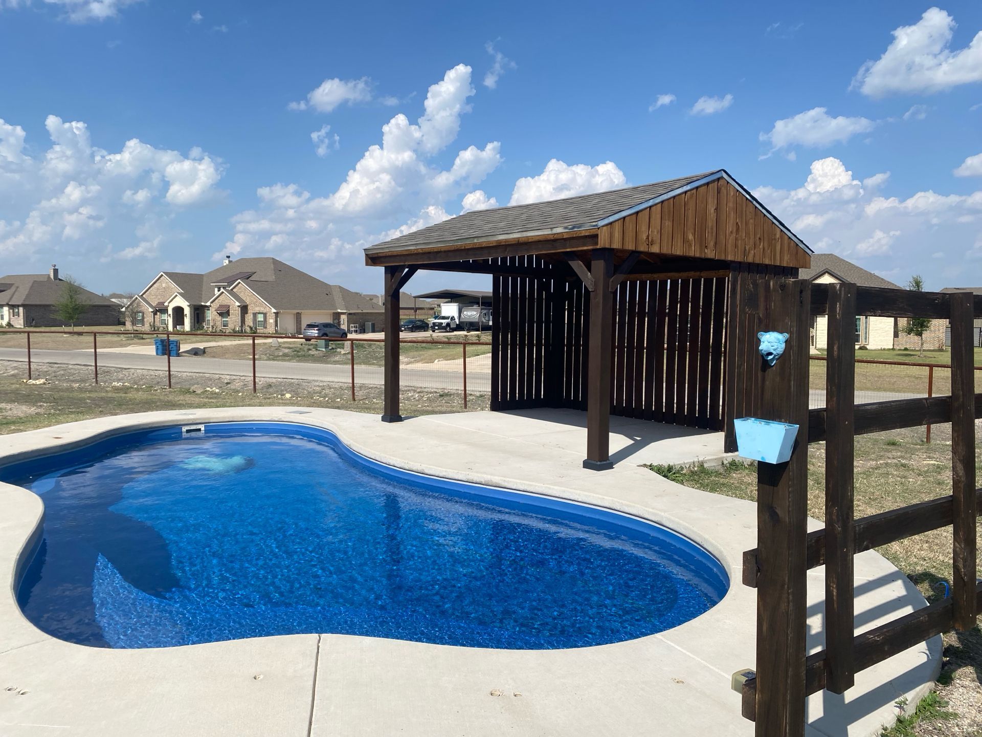 Poolside wooden cabana next to a blue, kidney-shaped swimming pool, with a fence, on a sunny day.