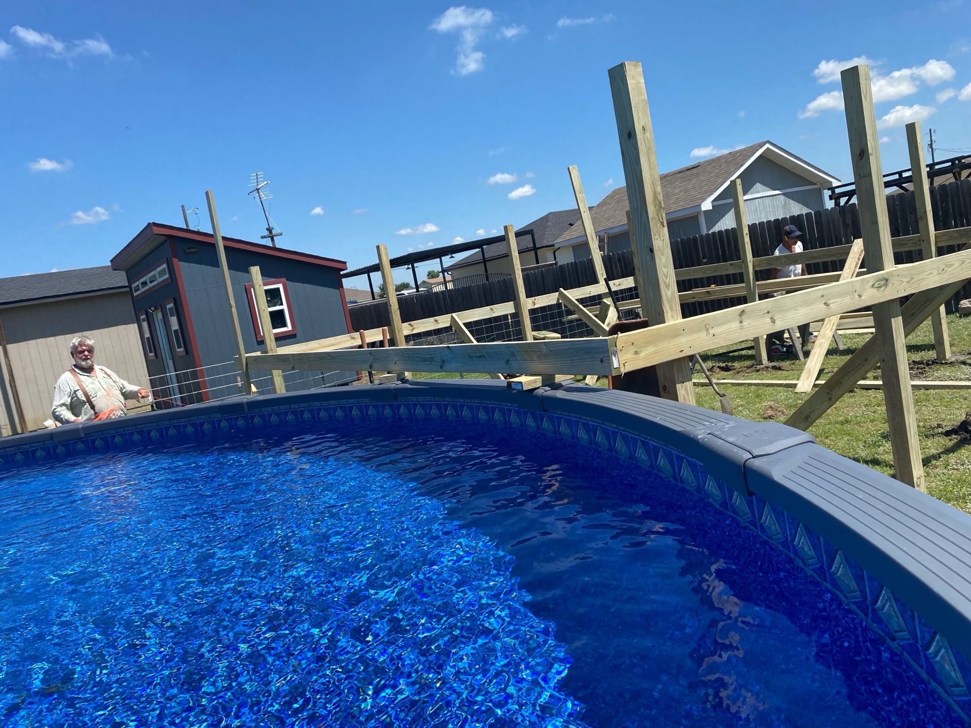A man near a partially built wooden deck next to a blue above-ground pool. The sky is clear and blue.