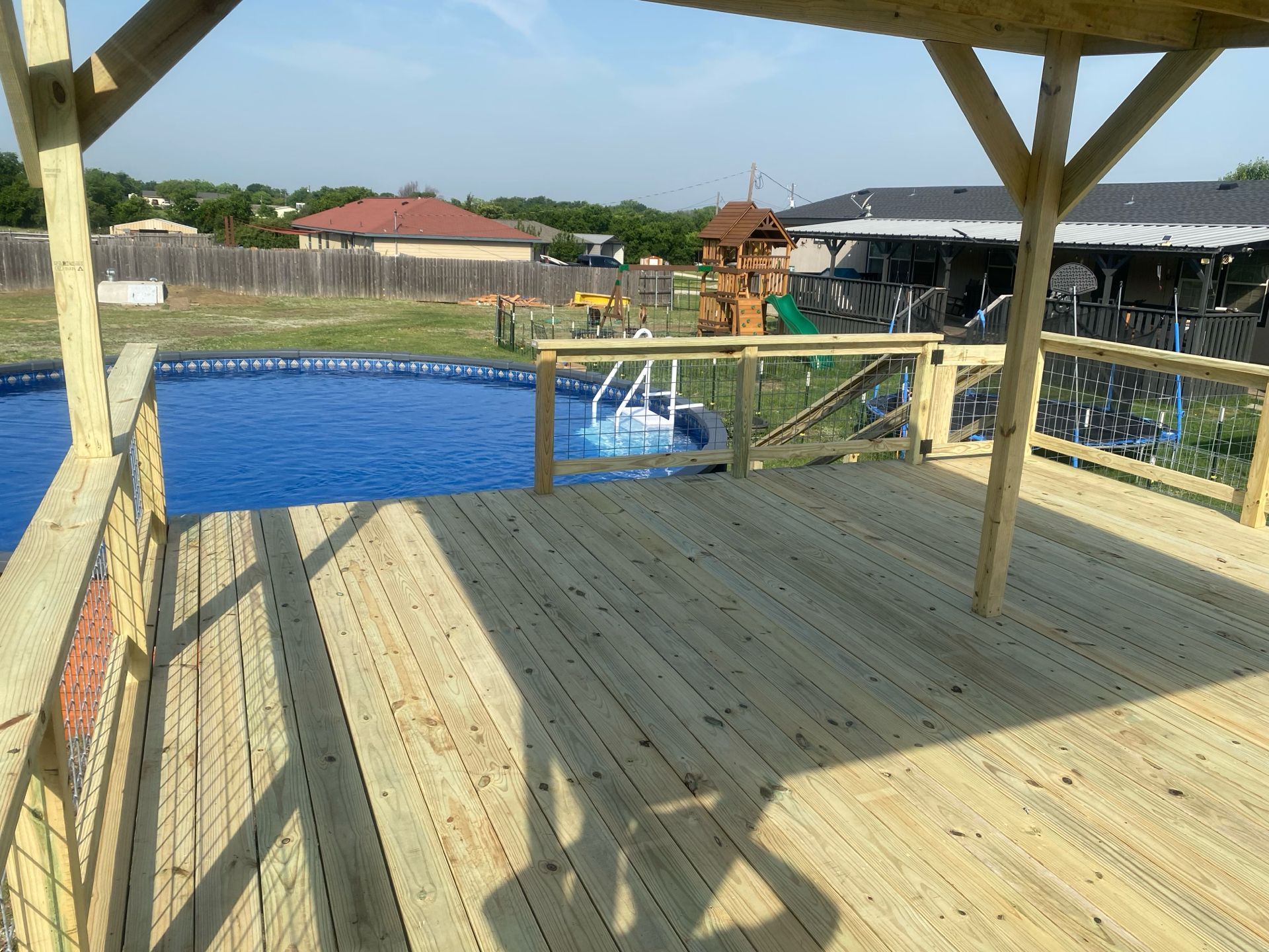 Wooden deck overlooking an above-ground pool and a playset in a backyard on a sunny day.