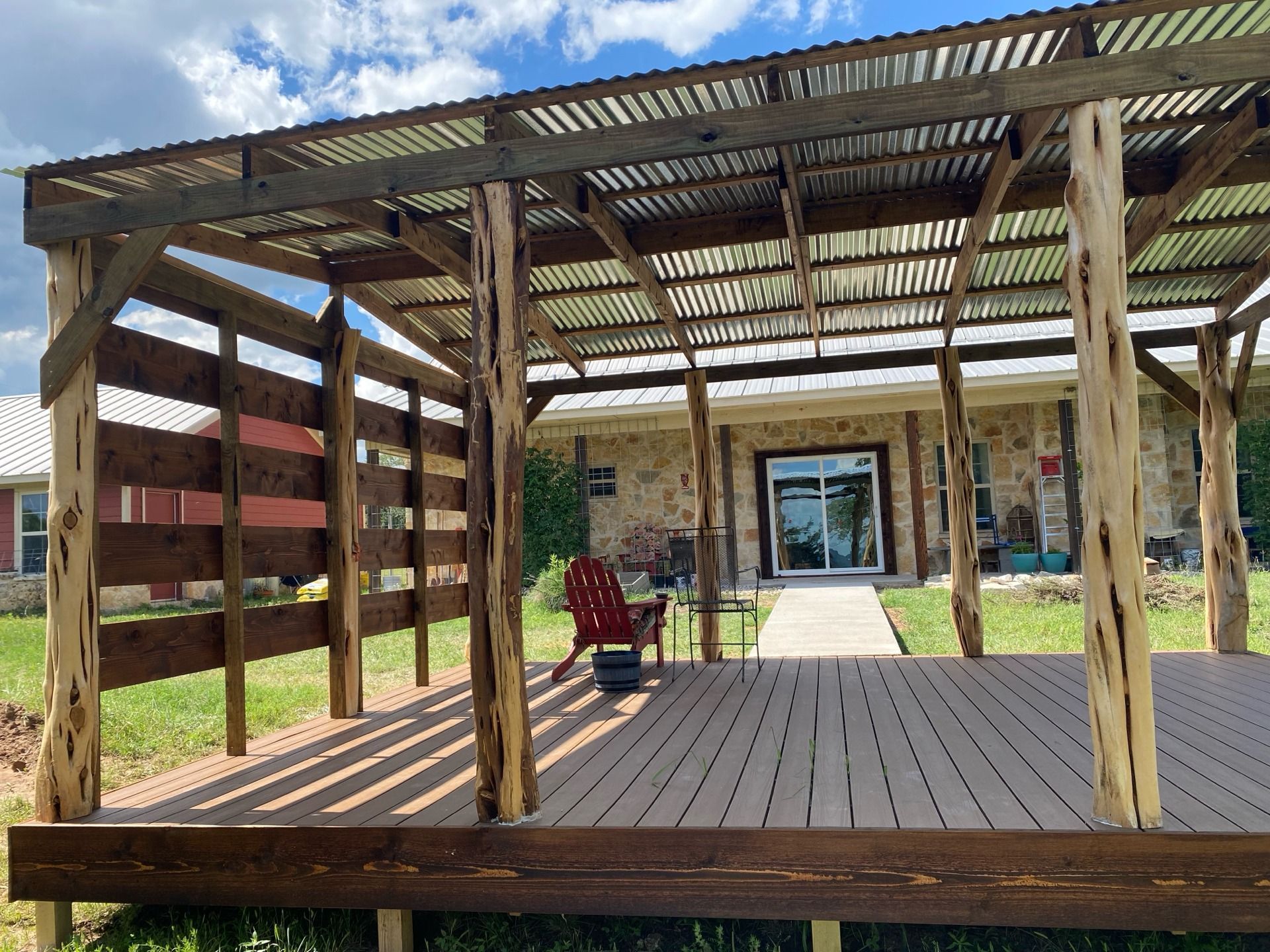 Wooden pergola with metal roof over a deck, red chair, and house with glass doors in the background.