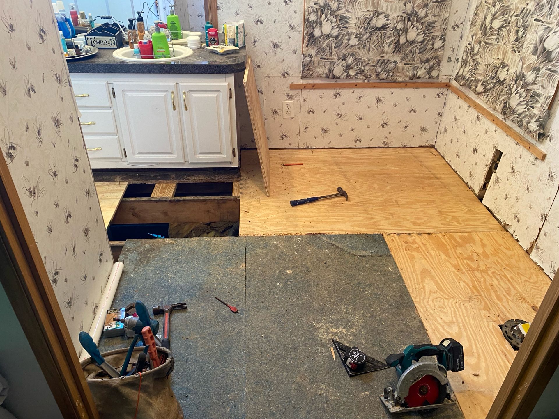 Bathroom under renovation: plywood flooring, exposed subfloor, tools scattered, sink with cabinet.