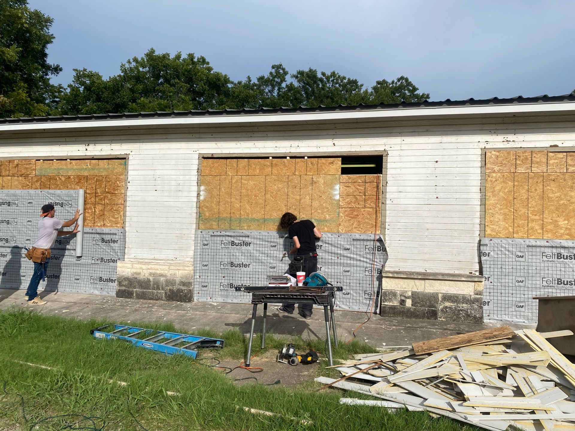 People installing siding on a white building with boarded-up windows; a table saw sits in front.
