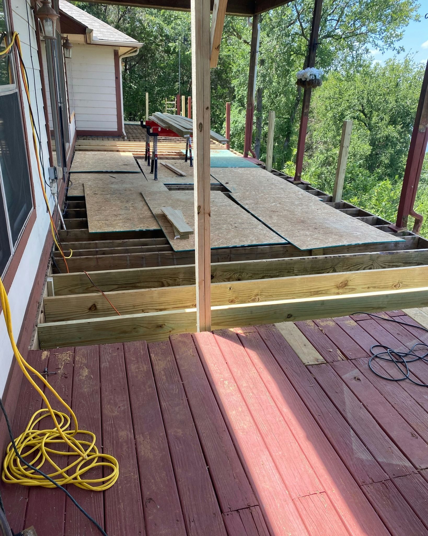 Deck construction in progress; weathered red deck, unfinished steps, partially boarded platform, green trees.