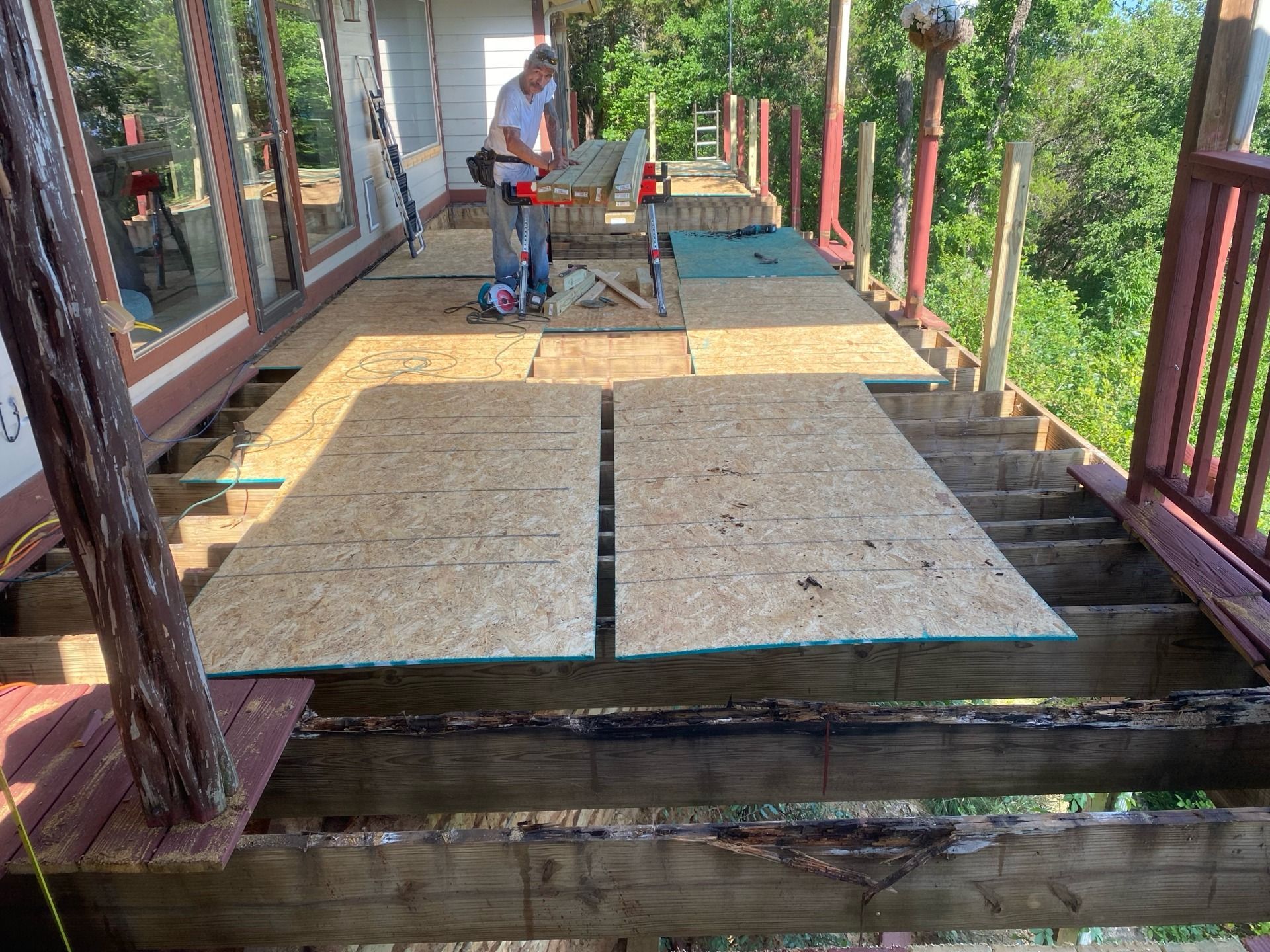 Person cutting plywood on a partially constructed wooden deck outdoors, surrounded by trees.